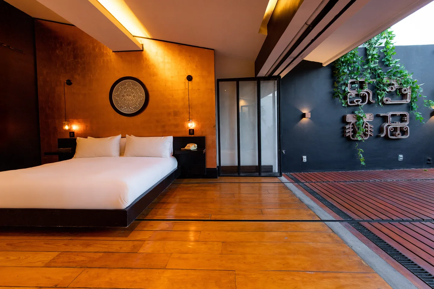 Bed against a copper-leaf wall with the glass doors open to the terrace at dusk — Polaris Suite, La Valise Mexico City