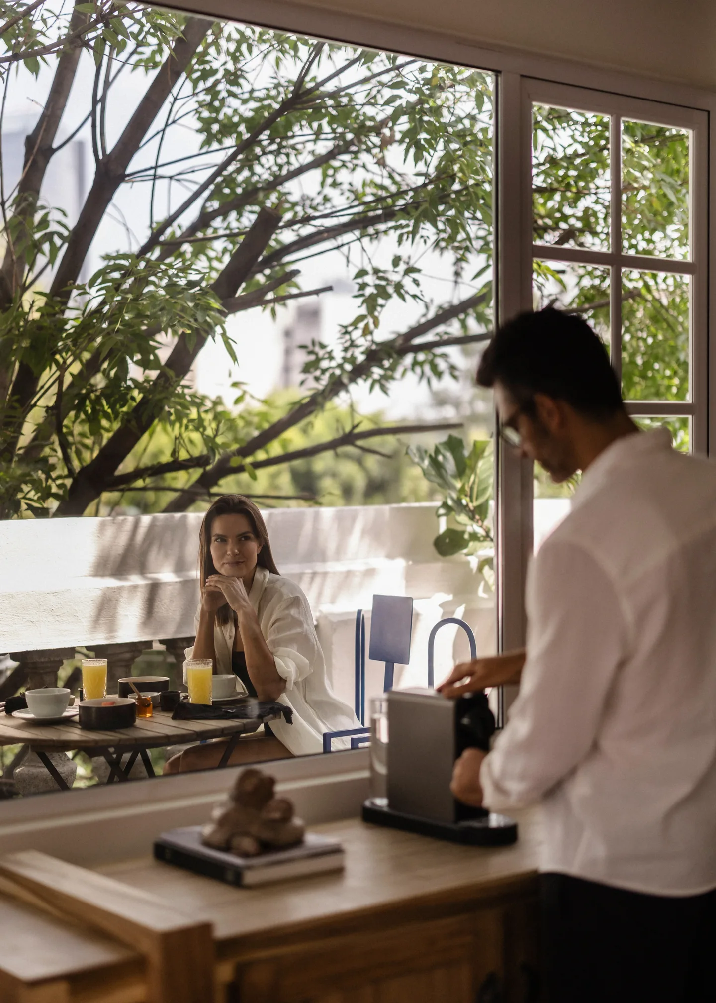 Host serving breakfast to a guest at a window table overlooking the trees of Roma Norte